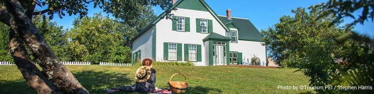 A little girls with red hair wearing 1800s period dress sits on a picnic blanket in front of a white house with green gables.